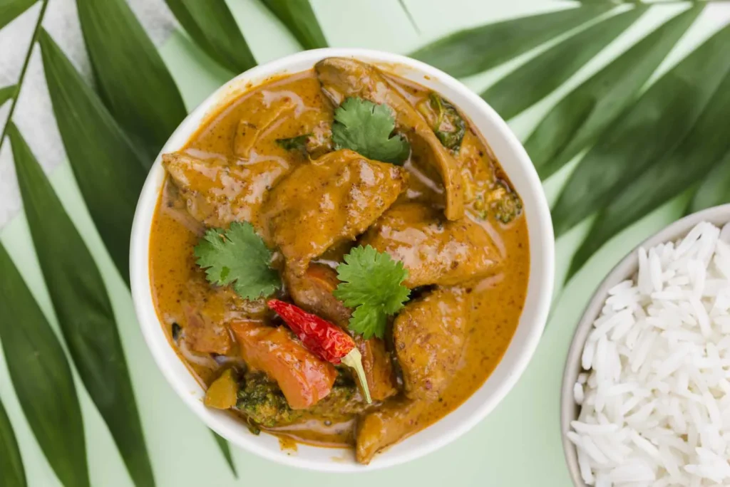 Curry de porc avec légumes, coriandre et piment dans un bol blanc, accompagnée de riz blanc, sur fond vert tropical.