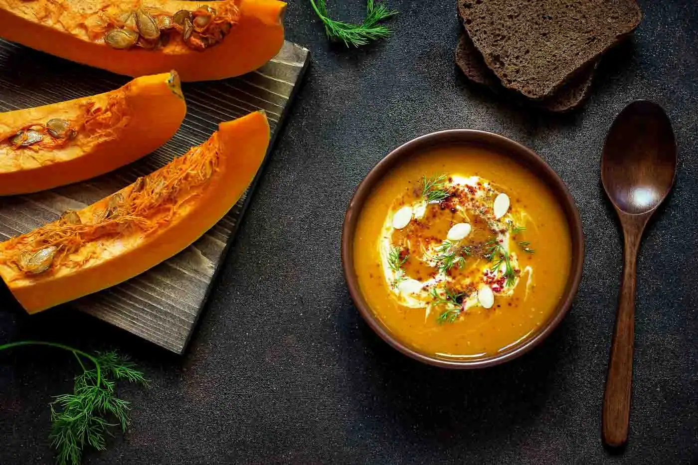 Velouté de potiron au curry à l'ancienne Velouté de potiron dans un bol, avec crème, herbes et épices, sur fond noir avec morceaux de potiron et cuillère en bois.