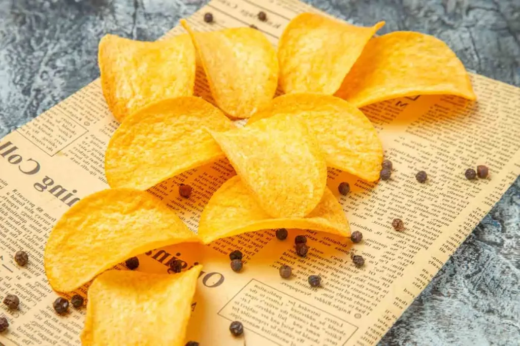 Chips dorées sur papier journal avec grains de sel et poivre noir, fond en pierre grise.