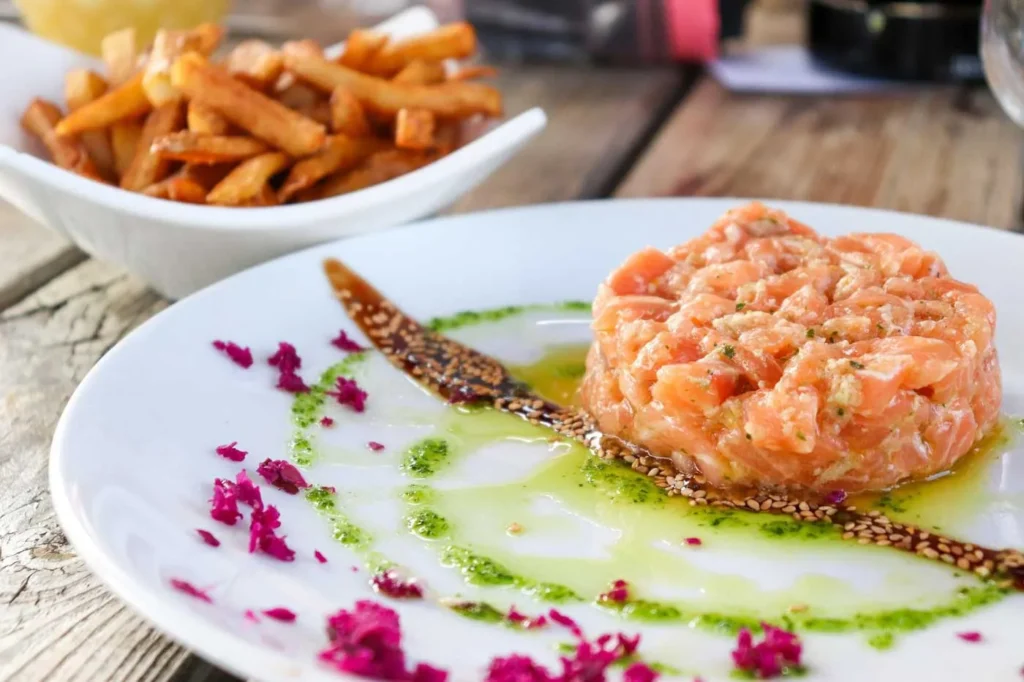 Tartare de saumon cru avec herbes, présenté avec frites dorées sur une assiette blanche, décorée de sauces vertes et roses.