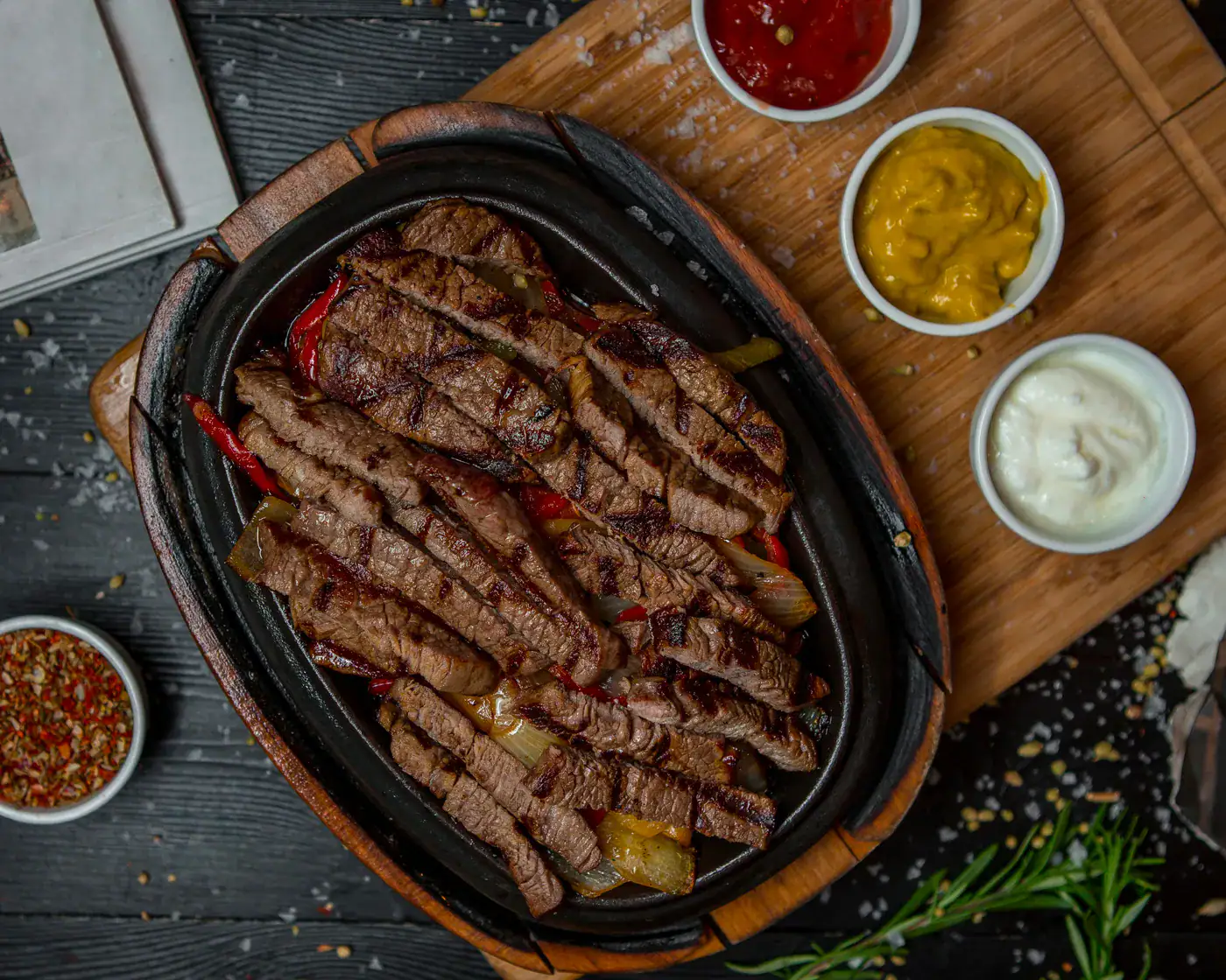 Boeuf grillé en tranches sur un plat en fonte, accompagnée de sauces moutarde, mayonnaise et épices en petits bols.