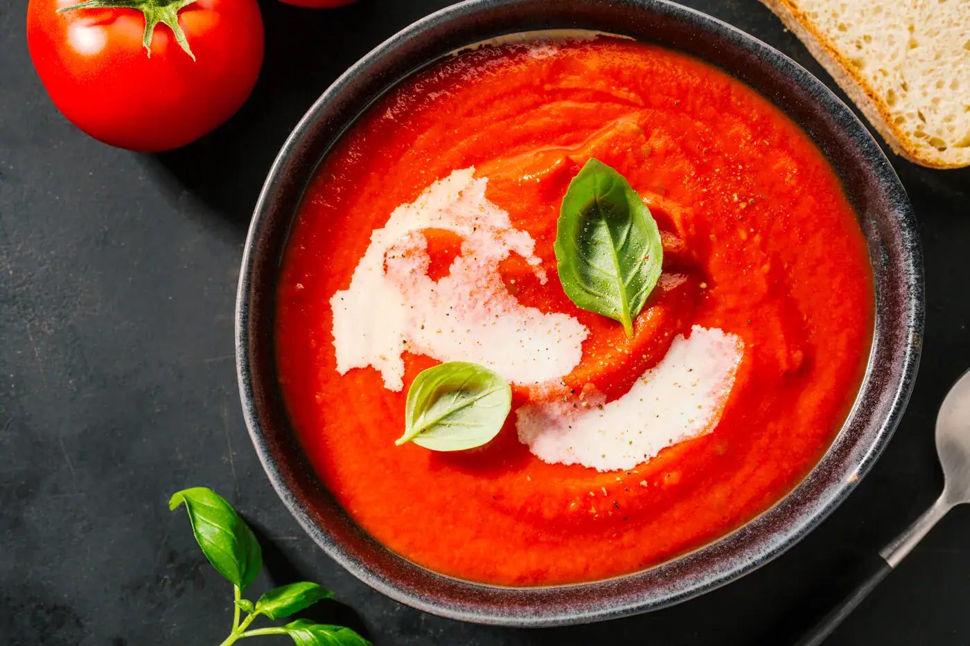 Gaspacho au Sel au Piment d'Espelette Soupe de tomate dans un bol noir, garnie de crème et de basilic frais, avec une tomate et du pain en arrière-plan.