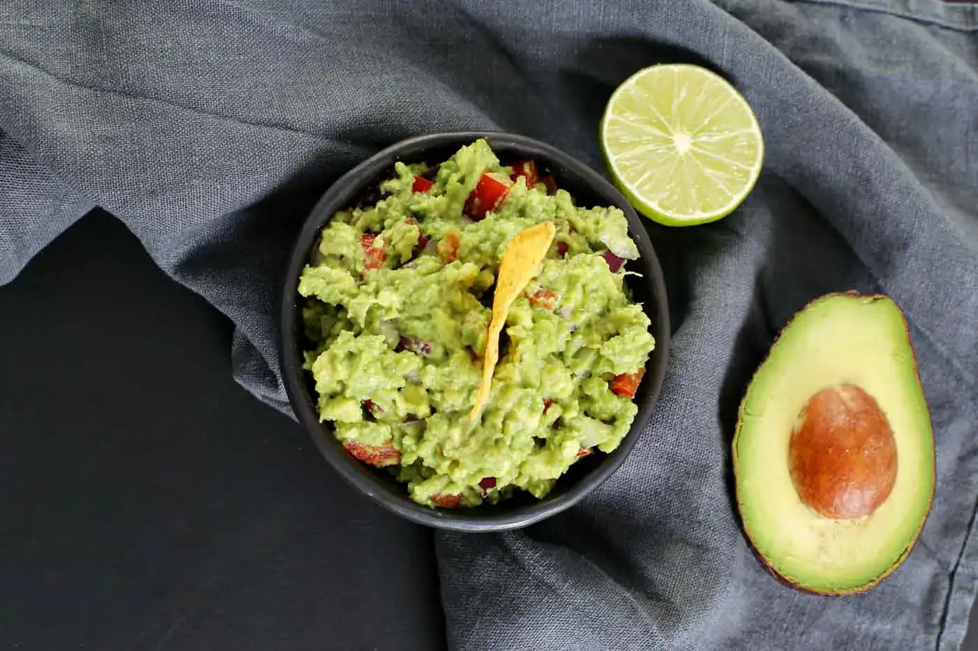 Guacamole dans un bol noir avec avocat coupé, citron vert et tranche de citron sur fond gris et lin.