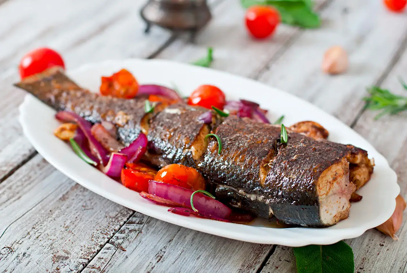 Poisson Grillé au Poivre Fumé de Tellicherry Sardines grillées sur une assiette avec tomates cerises, oignons rouges et herbes, sur fond en bois clair.