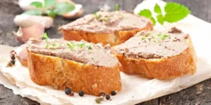 Terrine de foie gras aux morilles et quatre épices Tranches de terrine de foie gras avec poivre noir et herbes, sur papier parchemin, fond en bois clair.