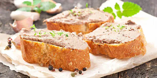 Tranches de terrine de foie gras avec poivre noir et herbes, sur papier parchemin, fond en bois clair.