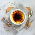 Crème brûlée dans un ramequin blanc, caramelisé en surface, sur un linge en lin gris clair, avec cuillère en métal à côté.