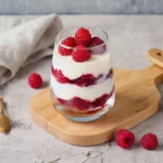 Verrine transparente avec couches de mascarpone et framboises fraîches, décorée de framboises entières, sur planche en bois.