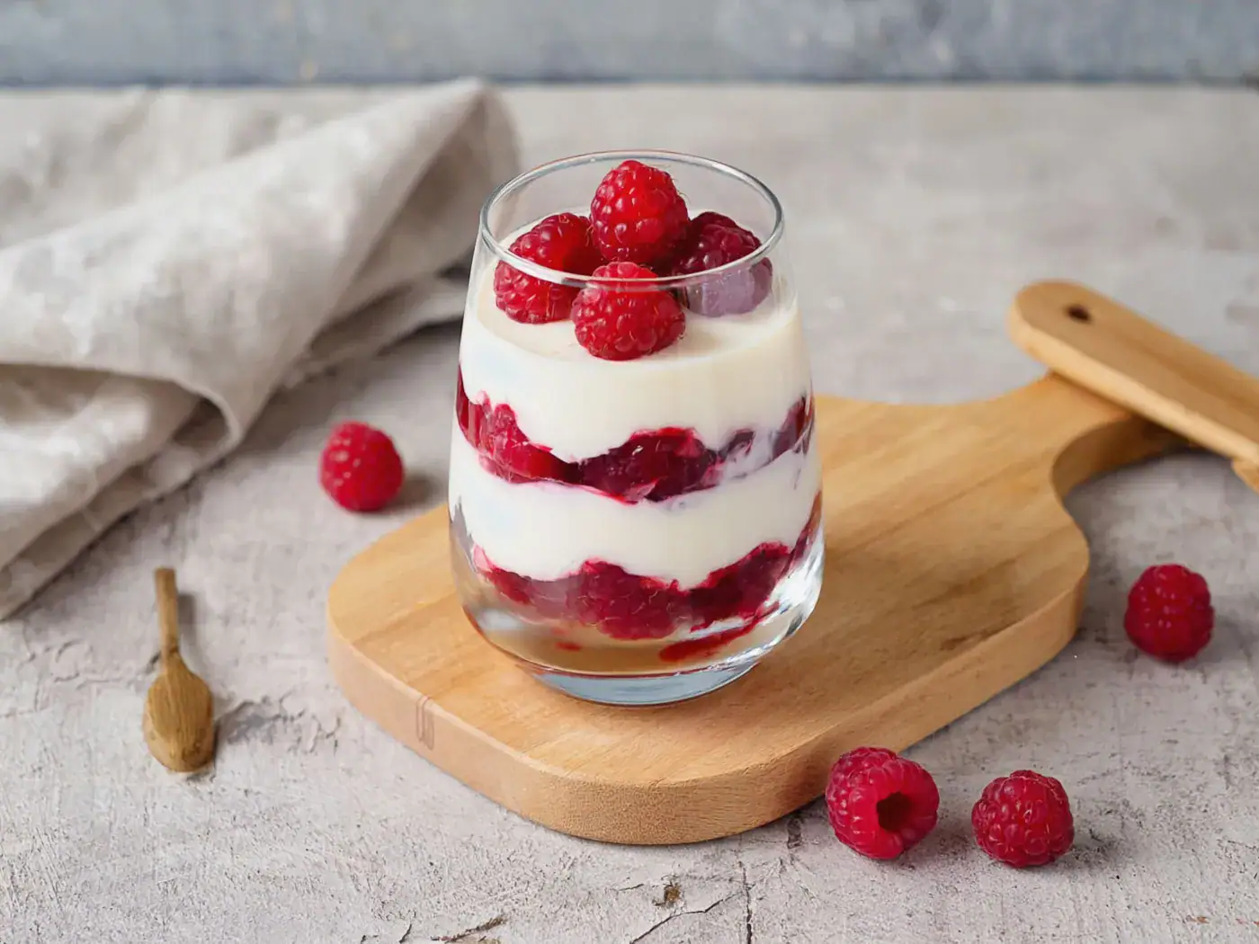 Verrine transparente avec couches de mascarpone et framboises fraîches, décorée de framboises entières, sur planche en bois.